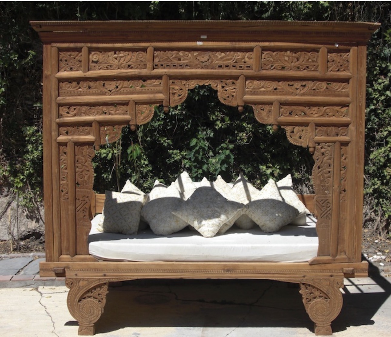 Cama-tallada de madera antigua con cojines blancos y grises en jardín