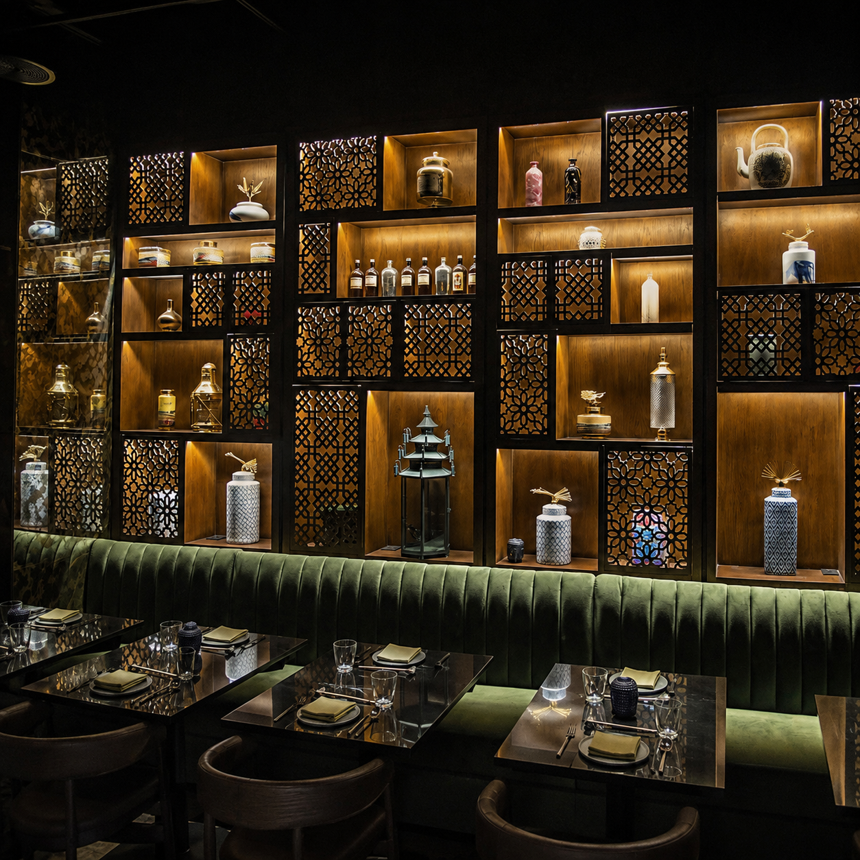 Bar de hotel con banquetas de terciopelo verde, mesas negras y estantería iluminada con cerámica